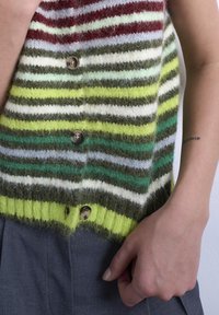 Κοντινό πλάν ο ενός ατόμου που φοράει ριγέ, μαλακό πλεκτό γιλέκο με κουμπιά, σκουρόχρωμο παντελόνι και έχει ένα μικρό τατουάζ εμφανές στο αντιβράχιο.