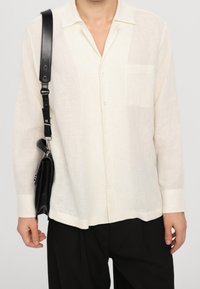 Camicia di lino beige chiaro con taschino, chiusura con bottoni e maniche lunghe. Indossata con una borsa nera con tracolla.