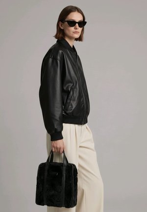 Mujer que lleva una chaqueta de cuero negra y gafas de sol, sosteniendo un bolso negro peludo, vestida con pantalones blancos y holgados frente a un fondo liso.