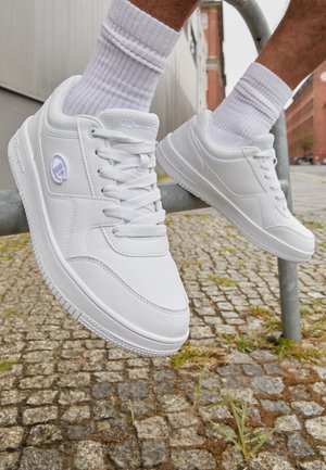 White sneakers with a smooth synthetic upper, lace-up design, padded collar, and logo detail on the side, against a cobblestone background.