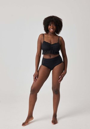 Glimlachende vrouw met krullend haar staat blootsvoets, gekleed in een zwart kanten crop top en hoog uitgesneden ondergoed, tegen een effen lichte achtergrond.