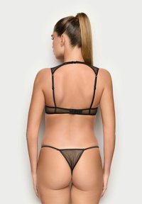 Donna con una lunga coda di cavallo che indossa un set di lingerie in rete nera, vista di spalle su uno sfondo chiaro e semplice.