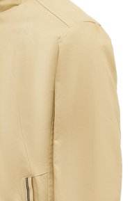 Beige letvægtsjakke med en glat tekstur, der har en høj krave, tydeligt synlige sømme og en skrå lomme med sort trim.