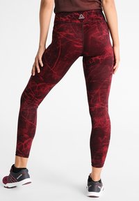 Leggings rossi a fantasia realizzati in materiale elasticizzato, con una vita alta e un logo Reebok sul retro, abbinati a scarpe da ginnastica nere.