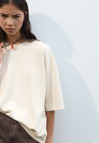 Cremfarbiges Oversize-T-Shirt mit kurzen Ärmeln und rundem Ausschnitt, kombiniert mit braunen strukturierten Hosen. Abgerundet mit einer klobigen Halskette.