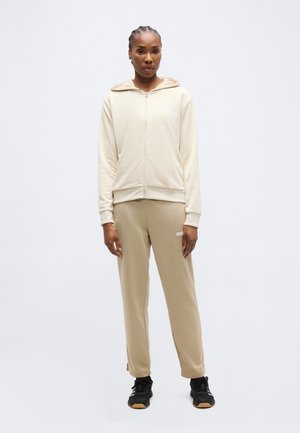 Femeie în picioare, purtând hanorac bej cu fermoar și pantaloni asortați, cu pantofi sport negri, pe un fundal simplu gri deschis.