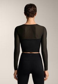 Fekete hosszú ujjú crop top áttetsző hátrésszel, sima textúrával és testhezálló kialakítással, magas derekú fekete leggingsszel párosítva.