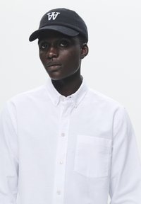 Camisa blanca de botones con cuello y bolsillo en el pecho izquierdo. Gorra de béisbol negra con el logo blanco de la "W". Diseño simple y limpio, con detalles mínimos.