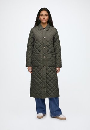 Femme portant un long manteau matelassé vert foncé avec des boutons, debout droite, associée à un jean bleu et des chaussures blanches sur fond blanc.