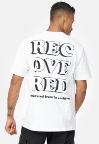 Hvid bomulds-T-shirt med fed sort tekst på ryggen: "GENVUNDET RØD," med "genvundet fra arkiverne" nedenfor i en mindre skrifttype.