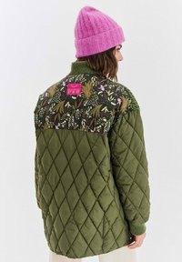 Prošívaná olivově zelená bunda s květinovým vzorem na horní části zad, s růžovou nášivkou a žebrovanými zelenými manžetami. Na hlavě vlněná růžová čepice.