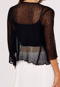 Czarny przejrzysty kardigan z długimi rękawami i falowanym wykończeniem na dole. Teksturowana tkanina z delikatnym wzorem, warstwowo ułożona na jaśniejszej bluzce.