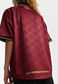 Persoon draagt een donkerrode sporttrui met subtiele geometrische patronen en gouden "INTERNATIONALS" tekst nabij de zoom aan de achterkant.