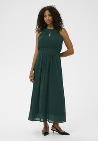 Rochie lungă, fluidă, de culoare verde închis, cu gât halter, detaliu în formă de cheie și talie smockată. Are un material ușor și este de lungime ankle.