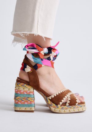 Sandalia de tacón alto de ante marrón con talón trenzado multicolor y envoltura de tela a rayas alrededor del tobillo, atada en un lazo, usada con pantalones blancos cortos.