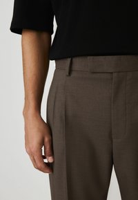 Braune maßgeschneiderte Hose mit glatter Textur, mit vorderen Falten und einem flachen Bund. Das Hemd ist schwarz mit kurzen Ärmeln.