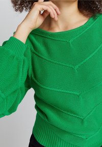 Zielony, dzianinowy sweter z teksturowanym wzorem chevron, łódkowym dekoltem i ściągaczami na rękawach. Materiał wydaje się miękki i swobodny.