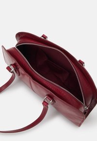 Sac à main en cuir rouge avec une fermeture éclair, une bandoulière plate et une texture lisse. L'intérieur comporte une poche unique et une doublure douce.