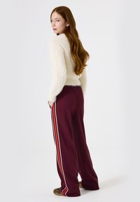 Suéter blanco esponjoso, pantalones burdeos con rayas blancas y naranjas a los lados, modelo con pelo largo mirando ligeramente hacia un lado, alejado de la cámara.