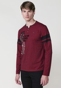 Hosszú ujjú burgundi ing texturált anyagból, henley nyakkal, fekete csíkozott részletekkel és fehér-fekete grafikákkal.