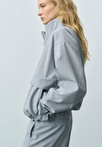 Ljust grå överdimensionerad jacka med hög krage, elastiska manschetter och en kort design. Tyget verkar slätt med en strukturerad silhuett.