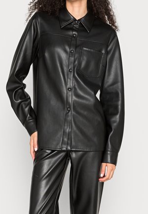 Frau trägt ein schwarzes Kunstleder-Hemd mit Brusttasche und passende schwarze Kunstlederhose, Hand auf der Hüfte.