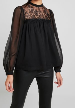 Žena v černé blůze s průsvitnými dlouhými rukávy a květinovou krajkovou vsadkou, doplněná černými koženými kalhotami na šedém pozadí.