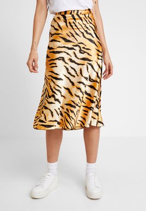 Femme portant une jupe en satin brillant avec des rayures tigre noires et orange, des baskets blanches et des chaussettes blanches sur un fond gris uni.