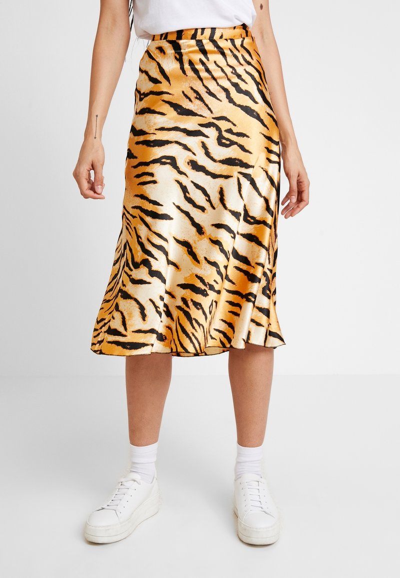 Femme portant une jupe en satin brillant avec des rayures tigre noires et orange, des baskets blanches et des chaussettes blanches sur un fond gris uni.