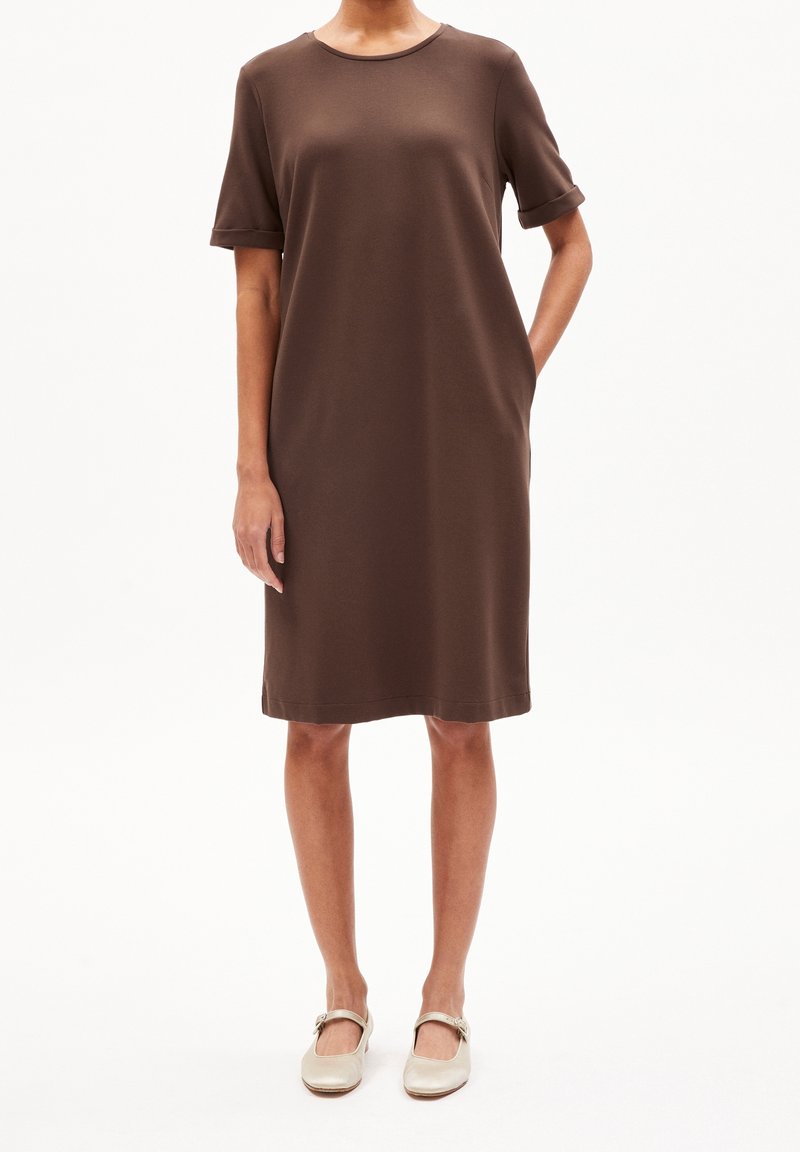 Femme portant une robe marron courte à manches courtes avec des poignets retroussés et des chaussures Mary Jane beiges, debout sur un fond blanc.
