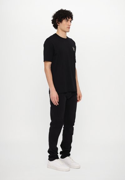 Juodas medvilninis marškinėliai trumpomis rankovėmis su maža logo ant krūtinės, derinami su juodomis kelnėmis ir baltais sportbačiais, stovintis ant vientiso fono.