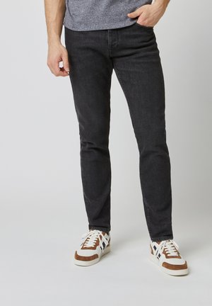 Jeans in denim nero con una vestibilità slim, caratterizzati da una leggera texture. Abbinati a sneakers marroni e bianche, che mostrano accenti in contrasto.