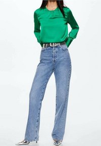 Grüne Satinbluse mit langen Ärmeln, kombiniert mit hochgeschnittenen blauen Jeans. Ein glänzender silberner Gürtel und spitze metallische Schuhe runden das Outfit ab.