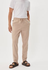 Helle beigen Drawstring-Hosen in lässigem Schnitt, aus einem weichen Stoff mit seitlichen Taschen, kombiniert mit braunen Slipper-Sandalen.