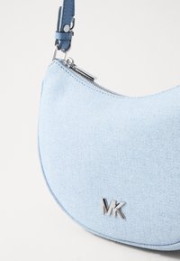 Džínsová kabelka v svetlo modrej farbe s zakriveným tvarom, s striebornými kovaniami a zipsom. Logo "MK" je výrazne zobrazené.