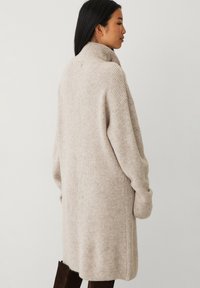 Beige gebreide cardigan met hoge kraag, oversized pasvorm, ribbeltextuur op de mouwen en zijzakken. Eenvoudig ontwerp, geen zichtbare patronen.