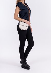 Borsa a tracolla bianca trapuntata con forma arrotondata, dotata di chiusura a pattina e tracolla regolabile marrone. Il modello indossa una blusa blu navy e jeans neri.