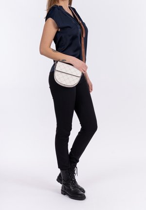 Bolso bandolera acolchado blanco con forma redondeada, que cuenta con un cierre de solapa y una correa ajustable marrón. La modelo lleva una blusa navy y jeans negros.