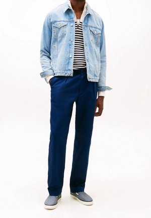 Mies, joka pit�� vaaleansinist�� farkkutakkia valkoisen ja mustan raidallisen paidan yli, tummansiniset housut ja harmaat liukkaat keng�t, joissa on valkoiset pohjat, seisoo k�det taskuissa.