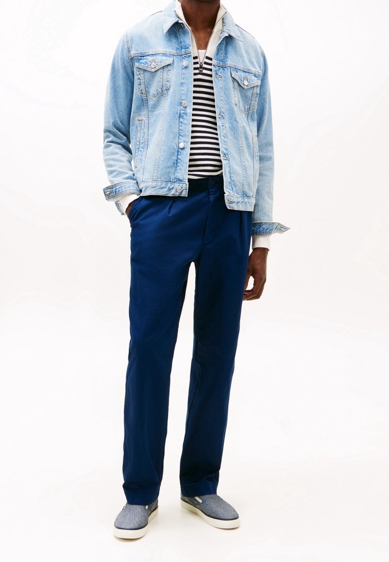 Jeansjacka, ljusblå, knäppt, med två bröstfickor. Randig skjorta under, marinblå byxor och gråa slip-on skor med vita sulor.