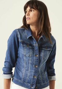 Denim jack in vervaagd blauw met knoopsluiting, dubbele borstzakken en opgerolde mouwen. Heeft een traditionele kraag en verticale naaddetails.