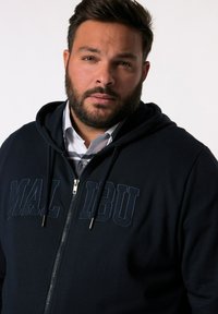 Marinblå zip-up huvtröja med stor "MALIBU" tryck i matchande färg. Har en dragsko i huva och silverfärgad dragkedja. Mjukt, texturerat tyg.