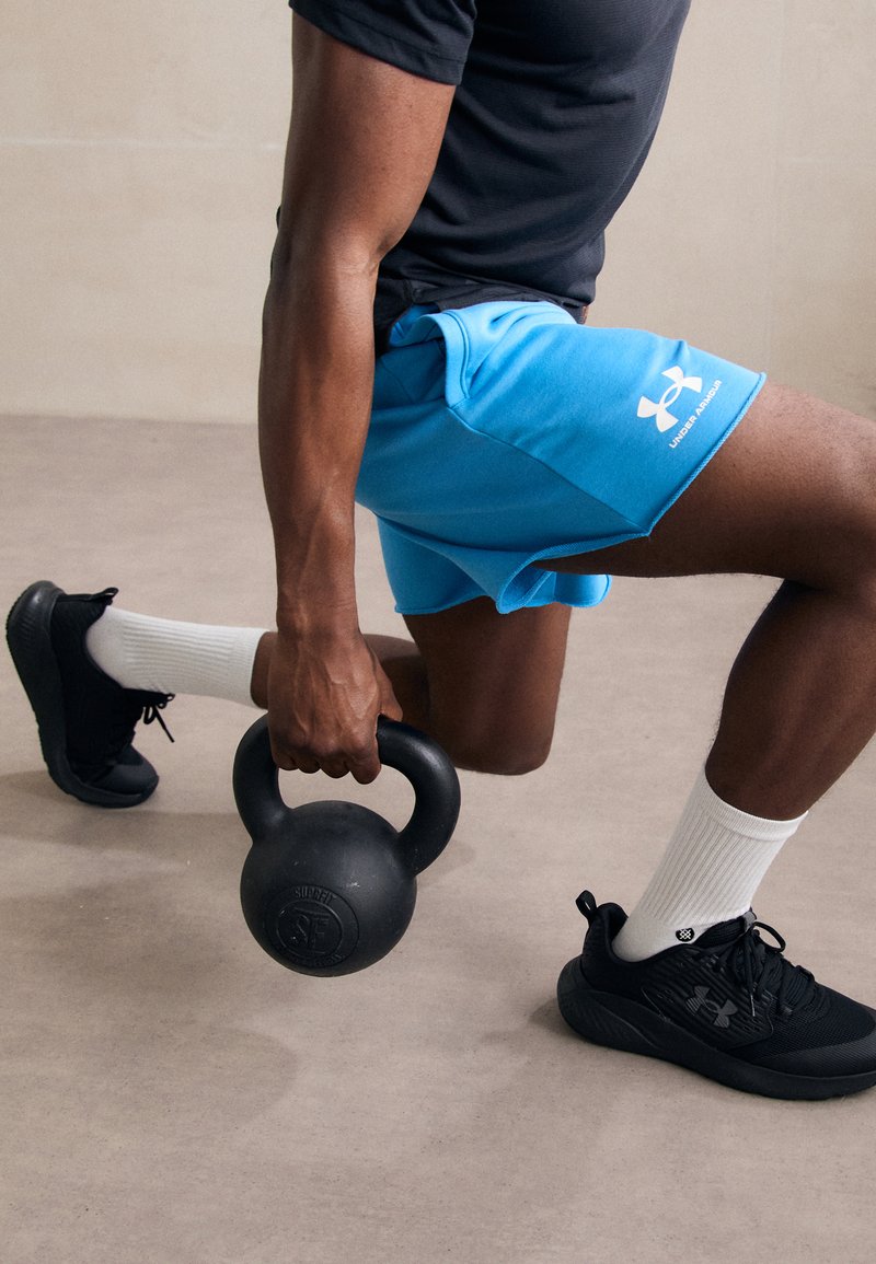 Schwarzes Kettlebell, gehalten von einer Person in einem schwarzen T-Shirt, türkisen Shorts und schwarzen Turnschuhen mit weißen Socken, positioniert auf einem hellgrauen Boden.