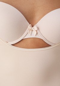 Beige molded bra with a smooth texture, featuring a cut-out design and a small bow accent at the center. Double-layered undergarment.