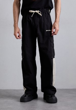 Man wearing black cargo pants with white side stripes, "Palm Angels" logo on thigh, white drawstring, and black platform shoes against plain background.
