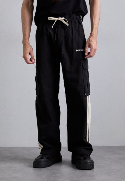 Man in zwarte cargopants met witte zijstreep, "Palm Angels" logo op de dij, witte trekkoord en zwarte platformschoenen tegen een effen achtergrond.