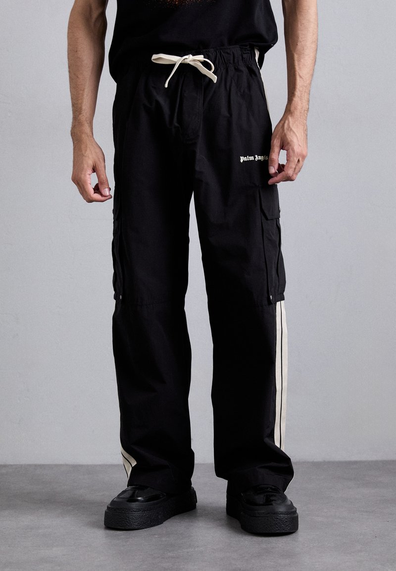 Man wearing black cargo pants with white side stripes, "Palm Angels" logo on thigh, white drawstring, and black platform shoes against plain background.