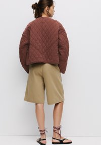 Jachetă quilted maronie cu o croială boxy, asortată cu pantaloni scurți bej până la genunchi și sandale negre cu șireturi înfășurate în jurul gleznelor.