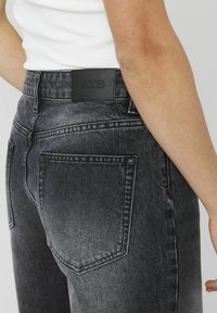 Jean en denim noir avec une finition délavée, comprenant deux poches arrière et un patch logo BZB sur la ceinture. Coupe ajustée avec des coutures visibles.