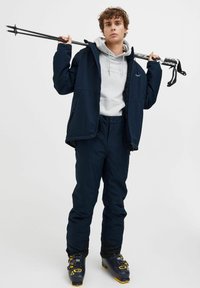 Costum de schi care include o jachetă albastră închisă și pantaloni asortați, un hanorac gri și bocanci de schi negri cu accente galbene. Ține în mână bețe de schi negre.